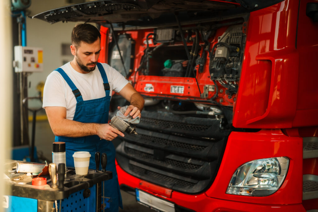 Mechanic in blue overalls works on a red truck engine in a garage, holding a tool. Nearby, a table holds various tools and a takeaway coffee cup.