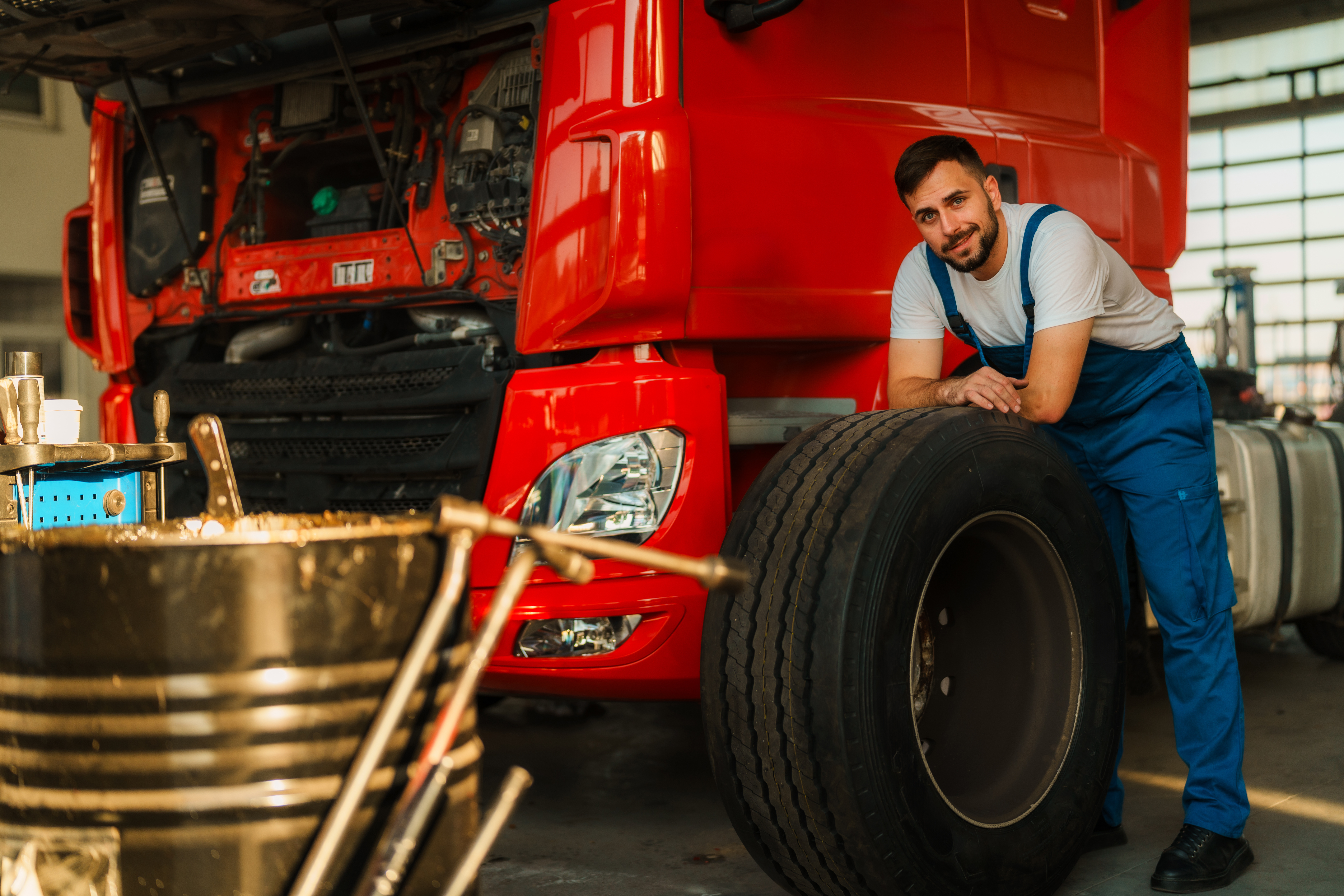 A man in blue overalls leans on a large tire beside a red truck with an open hood, surrounded by tools in a warm, well-lit garage. He smiles confidently.