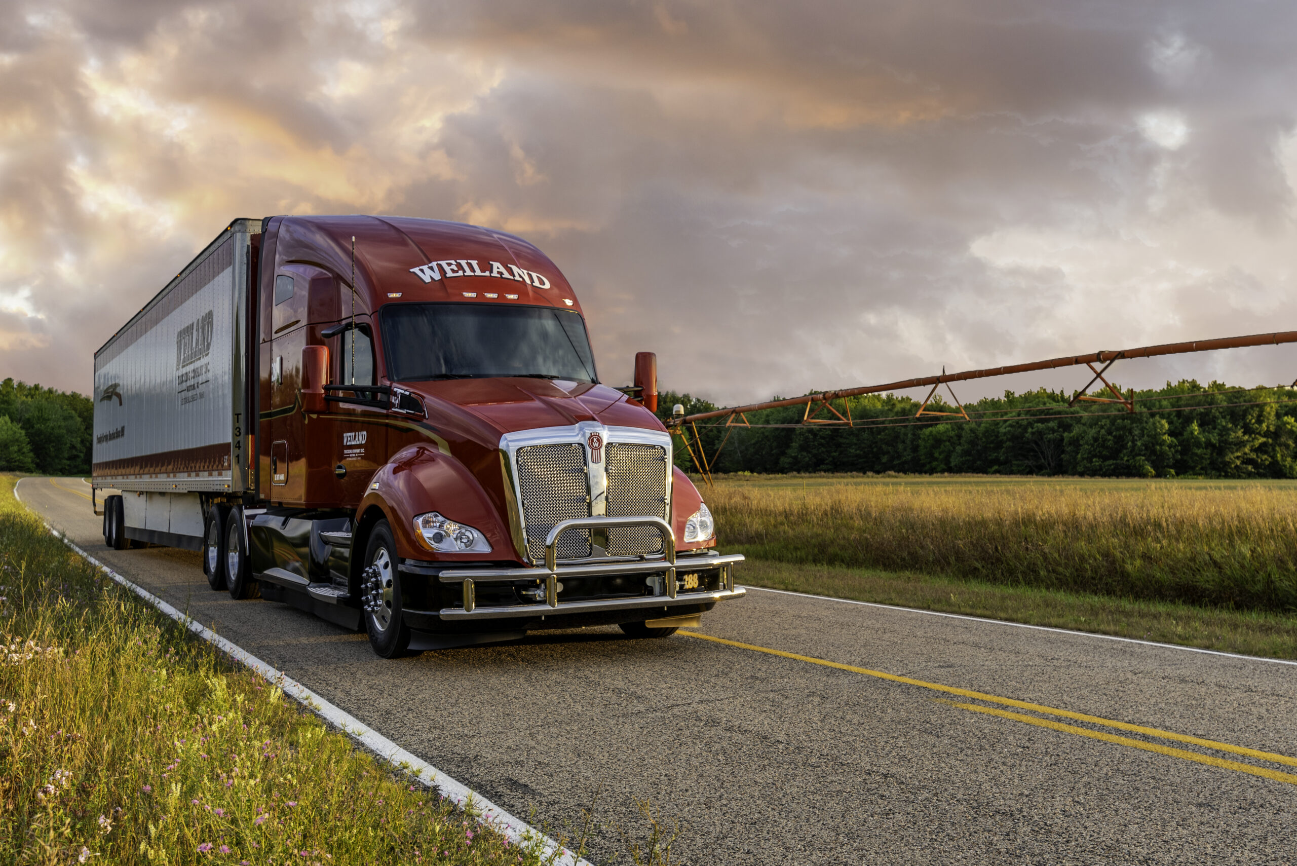 Red Weiland semi-truck driving on rural highway at sunset