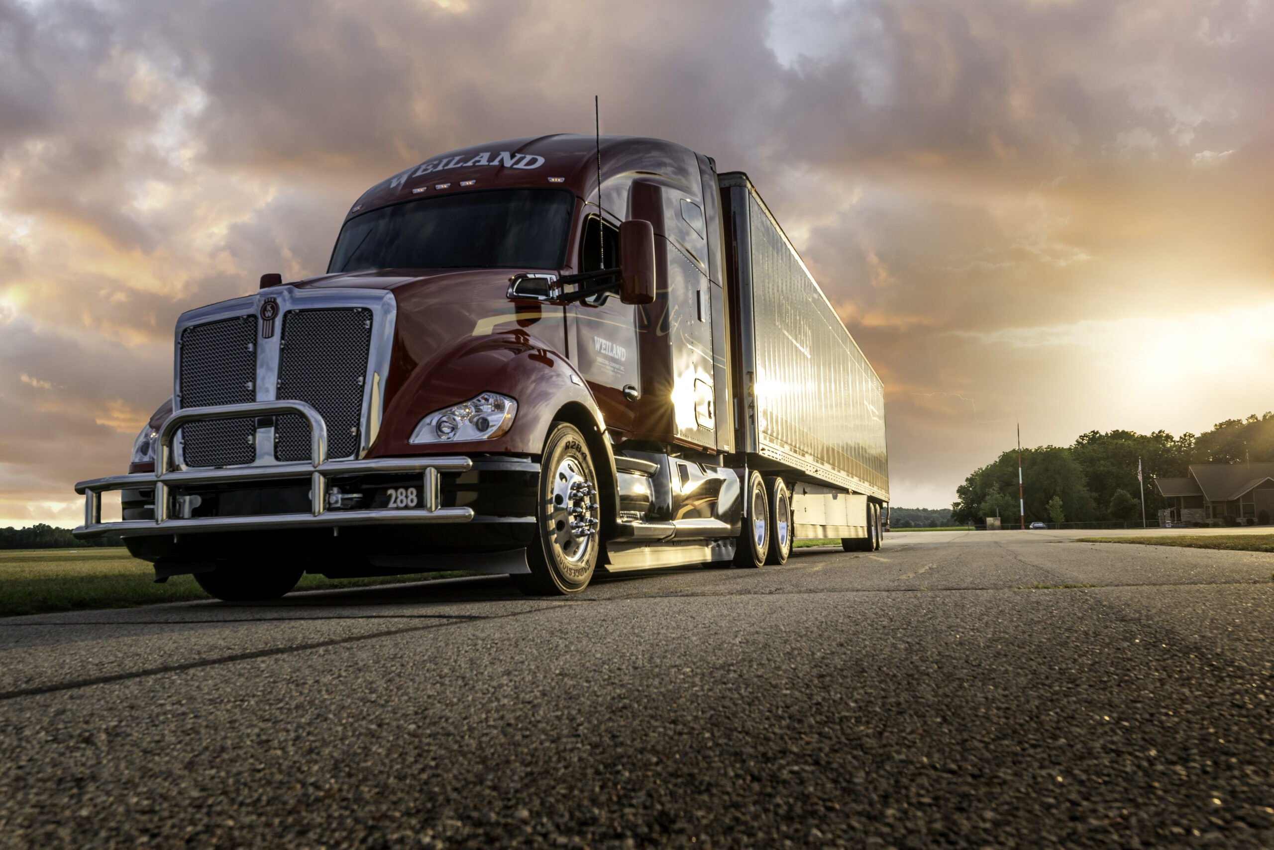 Red Weiland semi-truck with silver trailer on country road at sunset